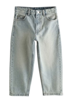 Jeune personne aux cheveux courts se tenant devant un fond beige, portant un t-shirt blanc, un jean bleu ample et des baskets noires, avec une main derrière la tête.
