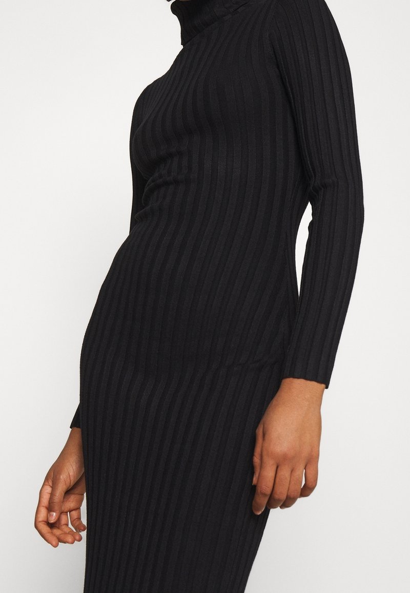 Personne portant une robe noire côtelée ajustée à manches longues avec un col roulé et des rayures verticales, visible du cou jusqu'aux genoux.