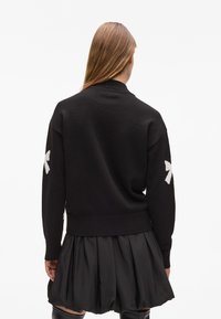 Czarny sweter z prążkowanym kołnierzem i mankietami, z białymi emblematami na rękawach. Materiał wydaje się być fakturowany i gruby.