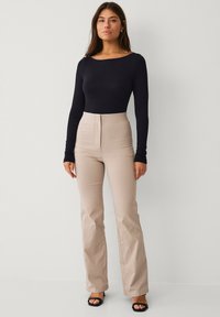 Schwarzes langärmeliges Bodysuit kombiniert mit beigen, hoch taillierten, ausgestellten Hosen, die eine glatte Textur, eine taillierte Passform und ein schlichtes Design aufweisen. Schwarze Sandalen.