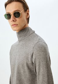 Grå polotröja i mjukt stickat tyg. Modellen bär runda solglasögon med guldinramning och mörka linser, med en neutral bakgrund.
