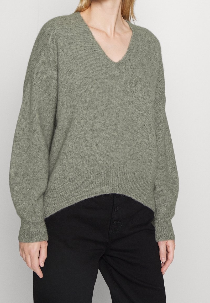 V-Ausschnitt gestrickter Pullover in Olivgrün, mit weit geschnittenen Ärmeln und einem gerippten Saum. Kombiniert mit hoch taillierten schwarzen Hosen.