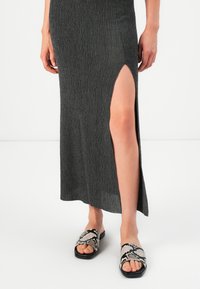 Ženska v strukturi temno sivi dolgi krili z stranskim razporkom in sandalih z odprto konico v kačjem vzorcu, stoji na beli tla.