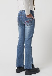 Jeans de mezclilla azul acampanados con bolsillos bordados, detalles de cremallera en el dobladillo y una cintura ajustada. Tejido texturizado con un lavado desgastado.