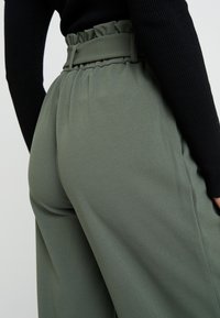 Zelené kalhoty s vysokým pasem vyrobené z hladké tkaniny, s volánkovým pasem a širokým střihem. Textura působí měkce a splývavě.