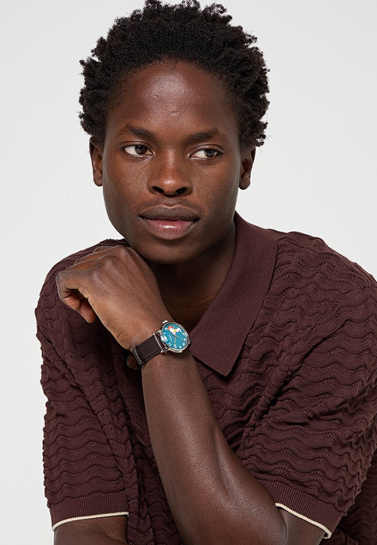 Hombre con cabello corto y rizado que lleva una camisa polo marrón texturizada y un reloj de pulsera con esfera azul y correa negra.