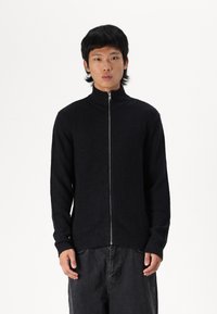 Maglione nero a coste con zip e colletto alto, dotato di maniche lunghe e zip in metallo, indossato con pantaloni scuri larghi.