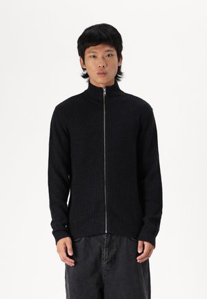 Suéter negro de canalé con cremallera y cuello alto, que presenta mangas largas y una cremallera de metal, combinado con pantalones oscuros de corte holgado.