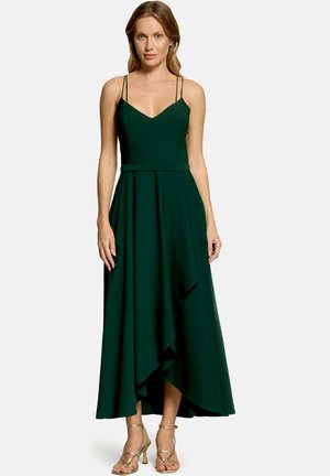 Femme portant une robe midi sans manches vert foncé avec de fines bretelles doubles et un ourlet asymétrique, associée à des sandales à talons hauts dorées à lanières.