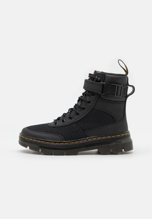 Bota alta negra con paneles de malla, cordones delanteros, correa ajustable con hebilla, lengüeta y suela gruesa ranurada con costuras amarillas.