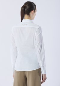Camicia bianca a maniche lunghe con colletto button-down, vestibilità su misura e delicato ricamo sul retro. Tessuto morbido e leggero.
