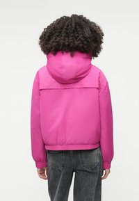 Jachetă cu glugă fuchsia, cu manșete și tiv elastic, realizată dintr-un material neted și ușor. Purtați peste blugi gri închis, văzută din spate.