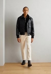 Chaqueta bomber de piel negra con dos bolsillos frontales, puños acanalados, llevada sobre una camiseta negra con cremallera. Pantalones de color beige claro y botas negras completan el look.