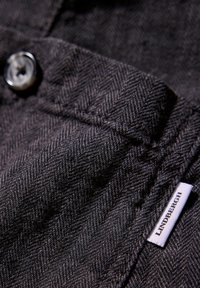 Närbild av svart fiskbensväv med en knapp och en vit etikett med texten "LINDENBERGH." Strukturerad yta med subtila mönsterdetaljer.