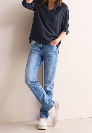 Kvinde iført mørkeblå langærmet top, lyseblå jeans og hvide sneakers, stående med den ene hånd i lommen og det ene ben krydset.
