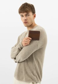 Brązowy skórzany portfel trzymany w dłoni, posiadający teksturowane wykończenie i prosty design. Noszony z jasnobeżowym swetrem z dzianiny.