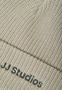 Beige strikket hue med en ribbet tekstur. Har en foldet kant med "JJ Studios" præget i sort på forsiden.