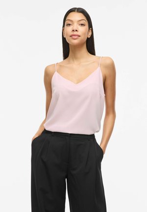 Kobieta w jasnoróżowej, bezrękawnikowej bluzce i czarnych spodniach z wysokim stanem, stojąca z rękami w kieszeniach na białym tle.