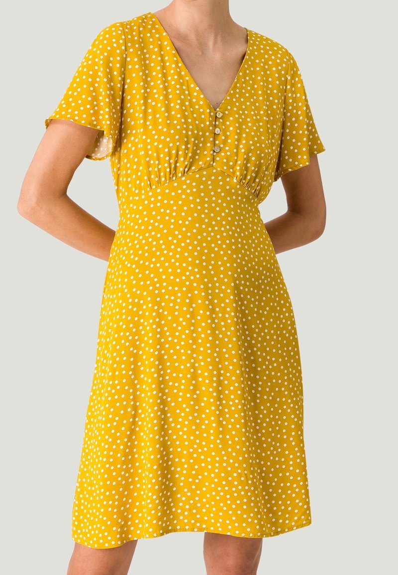 Femme portant une robe jaune mi-longue à pois blancs, manches courtes, col en V et trois boutons sur la poitrine, debout les mains derrière le dos.
