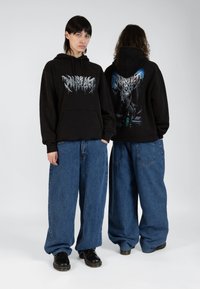 Black sweatshirt with front logo and back graphic design. Loose-fitting blue denim pants. Black shoes. Minimalist background.