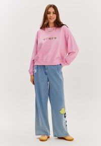 Růžová mikina s grafickým designem, volný střih a žebrované manžety, spojená se širokými světle modrými džíny s postavičkou z kresleného filmu.