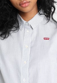 Camicia a maniche lunghe con bottoni, bianca con righe verticali blu navy, presenta un'etichetta con il logo Levi's sul petto sinistro e colletto standard.