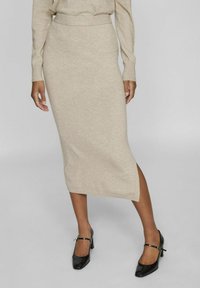 Falda midi de punto en color beige con cintura alta, abertura lateral y textura suave. Combinada con tacones de charol negros con tiras al tobillo.