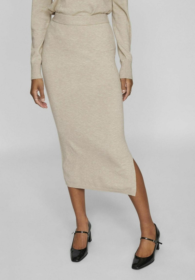 Falda midi de punto en color beige con cintura alta, abertura lateral y textura suave. Combinada con tacones de charol negros con tiras al tobillo.