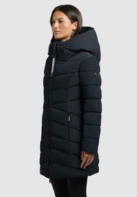 Marineblaue Steppjacke mit hohem Kragen, Kapuze und Zickzack-Steppmuster. Verfügt über einen Frontreißverschluss und Logo-Akzente am Reißverschluss und am Ärmel.