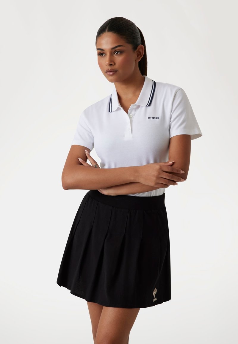 Weißes Poloshirt mit blau gestreiftem Kragen, mit 'GUESS'-Logo. Schwarzer Faltenrock mit elastischem Bund und kleinem goldenen Logo-Detail.