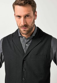 Schwarze Wollweste mit Reverskragen, vorderer Knopfleiste und Brusttasche, getragen über einem dunkelgrauen Hemd mit Kragen. Glatte Textur.