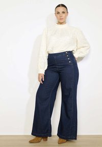 Pantaloni in denim blu scuro a vita alta con bottoni laterali, abbinati a una blusa testurizzata bianco sporco con maniche a sbuffo.