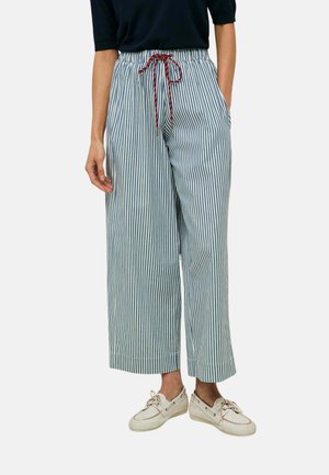 Mujer que lleva pantalones anchos de rayas verticales azules y blancas con cordón rojo, top negro y zapatos beige con cordones.