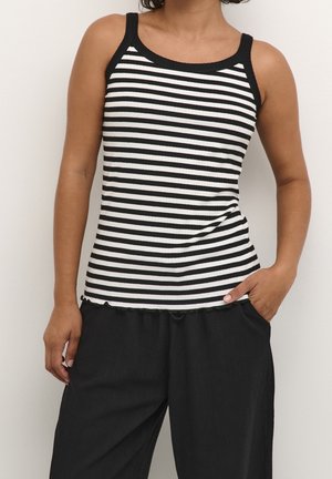 Kobieta ubrana w czarno-biały, poziomo paskowany top bez rękawów i luźne czarne spodnie, z jedną ręką w kieszeni na jednolitym tle.