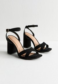 Sandalias de ante negro con tacón cuadrado y tiras cruzadas, y hebilla en el tobillo. Diseño de punta abierta y plantilla suave de color beige.