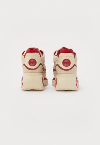 Zapatillas de color crema con acentos rojos, que cuentan con una suela gruesa, paneles texturizados y un parche circular con logo en el talón.