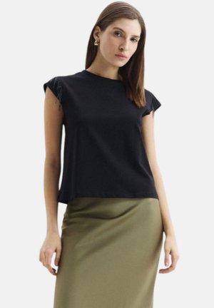 Donna che indossa un top nero senza maniche con spalle frangiate e una gonna verde oliva, in piedi contro uno sfondo bianco semplice.