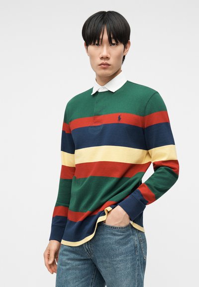 Νεαρός άνδρας που φοράει μακρυμάνικο ρούγκμπι με πράσινες, κόκκινες, ναυτικές και κίτρινες ρίγες, συνδυασμένο με μπλε τζιν, στέκεται με το ένα χέρι στην τσέπη.