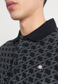Gros plan sur une personne portant un polo noir avec un motif de chaîne entrelacée grise et un petit logo triangulaire sur la poitrine.