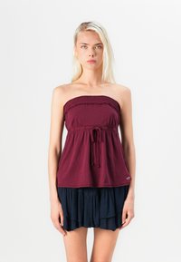Bordeaux strapless top met smockafwerking en verstelbare strik. Gepaard met een gelaagde marineblauwe rok met ruffles. Gladde textuur, casual ontwerp.