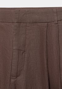 Pantalon en lin marron avec taille, passants de ceinture, plis avant et poches latérales visibles en gros plan.