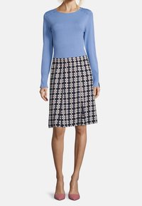 Vaaleansininen neulepaita, jossa on napit olkapäillä, yhdistetty laivastonsiniseen hameeseen, jossa on geometrinen vaaleanpunainen kuvio. Vaaleanpunaiset kengät viimeistelevät asun.