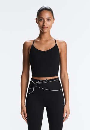 Frau steht frontal, trägt ein schwarzes kurzes Tanktop und schwarze Leggings mit hoher Taille und einem gebundenen Taillenband.