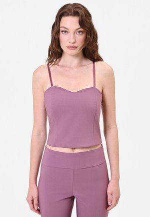 Giovane donna con capelli castani ondulati che indossa un crop top viola senza maniche e pantaloni a vita alta abbinati, in piedi davanti a uno sfondo bianco.