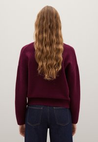 Burgunderfarbener Strickpullover mit lockerer Passform und geripptem Saum, kombiniert mit dunkelblauen Jeans; welliges Haar, das den Rücken hinunterfällt.