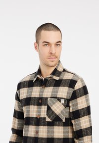 Geruite flanellen shirt in zwart, beige en grijs. Heeft een knoopsluiting aan de voorkant, een borstzak en houten knopen. Zachte textuur.