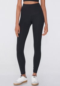 Leggings noirs taille haute en tissu extensible, avec une texture lisse et une coupe ajustée. Portés avec des baskets blanches.