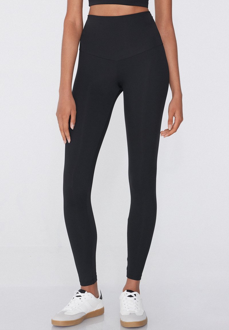 Leggings noirs taille haute en tissu extensible, avec une texture lisse et une coupe ajustée. Portés avec des baskets blanches.