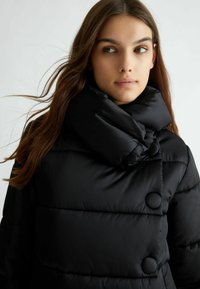 Cappotto puffe nero con colletto alto, grandi bottoni e texture trapuntata. Il tessuto appare liscio e ha una finitura lucida.