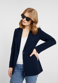Sacou marina cu un design croit, închidere cu un singur nasture și umeri ușor îmbățiți. Purtați peste un top alb și asortați cu blugi albaștri.